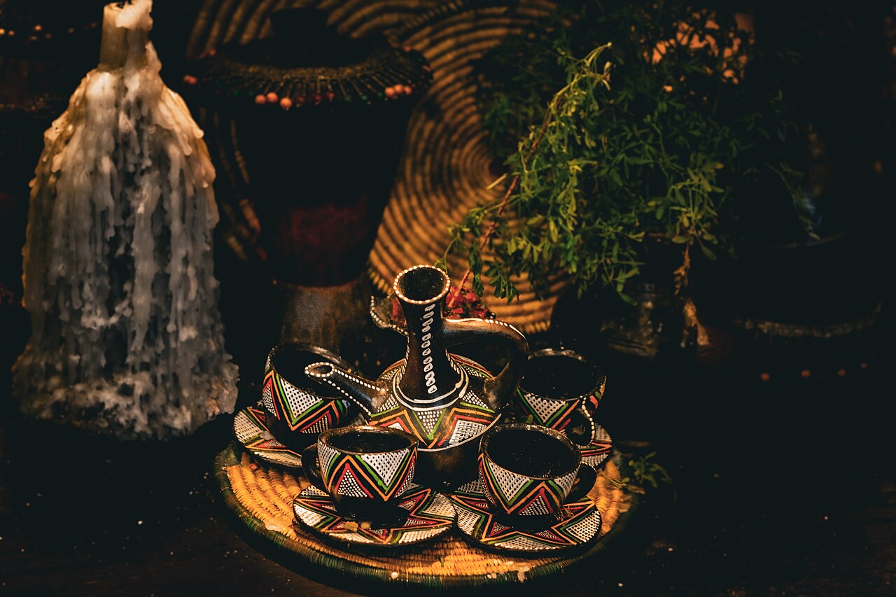 An Ethiopian woman wearing traditional dress, carefully pouring coffee from a jebena into small finjal cups as part of a coffee ceremony, with roasted beans and incense in the foreground.