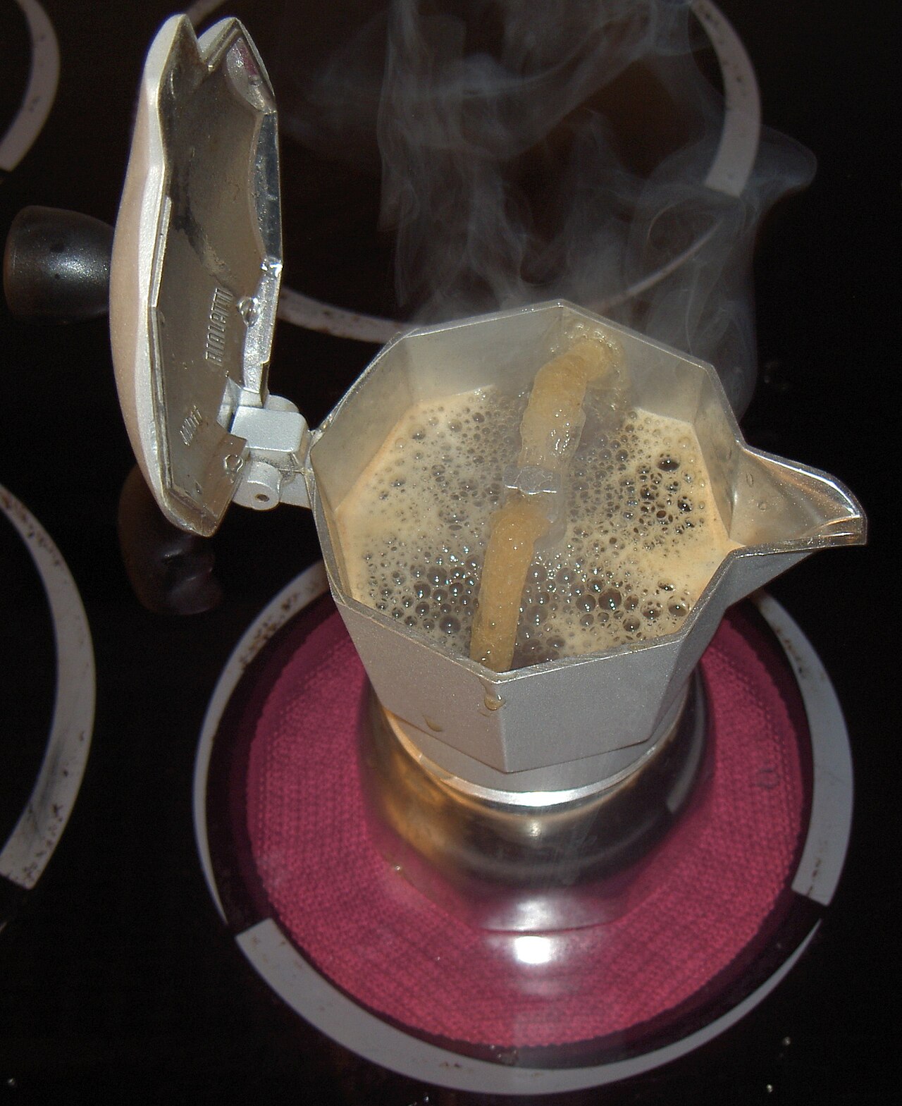 Close-up of a stainless steel Bialetti Moka pot brewing coffee on a gas stovetop, steam rising from the spout.