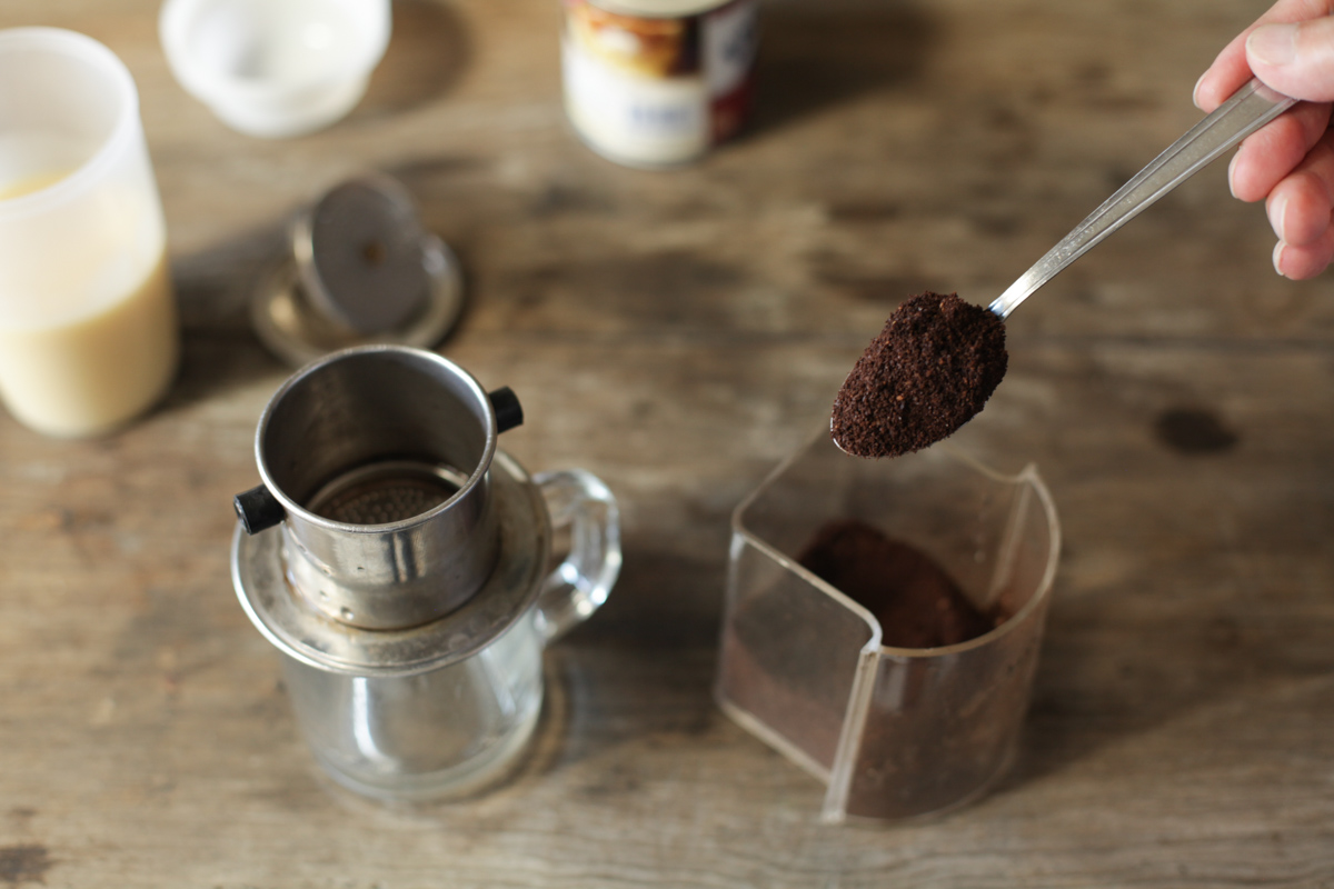 A stainless steel Vietnamese phin dripper slowly dripping coffee into a glass with sweetened condensed milk, with ice nearby.